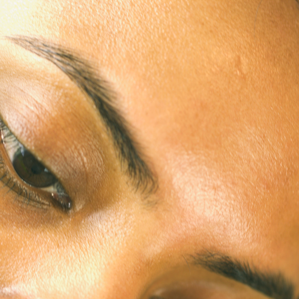 Close-up of a person's eye and eyebrow area, showing natural skin texture and dark brown eyebrow makeup.