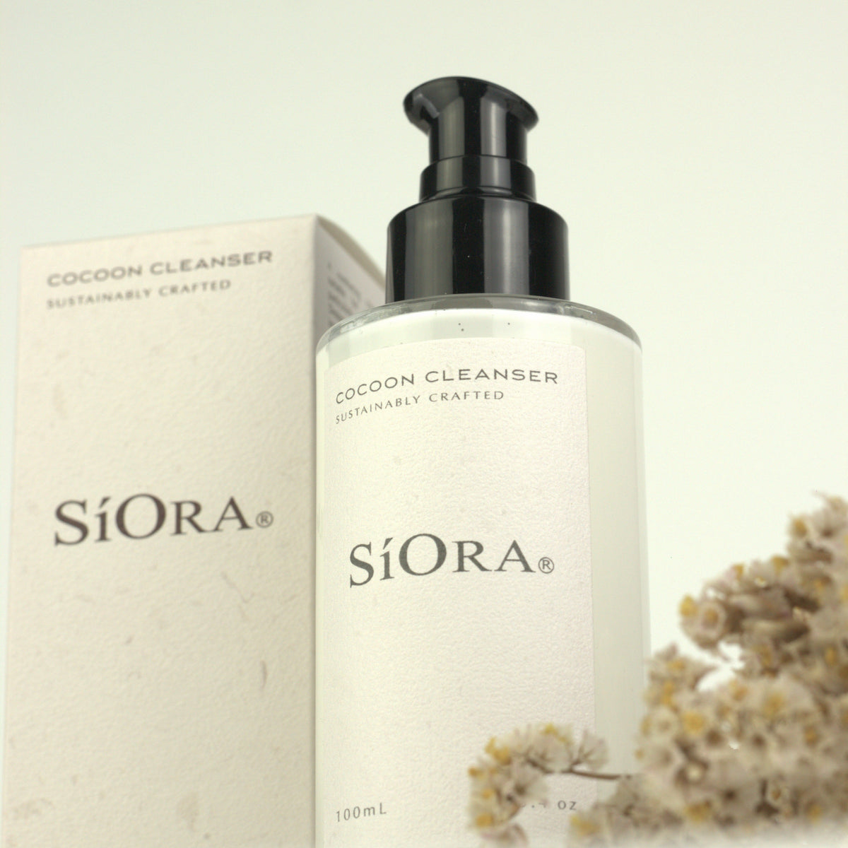 SiOra Cocoon Cleanser 100ml bottle with a black pump dispenser next to its textured off-white packaging box, styled with dried flowers on a light beige background.
