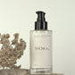 SiORA Cocoon Cleanser bottle with a black pump and clear glass body, displayed on a wooden block next to dried flowers and a patterned cloth against a warm cream background.