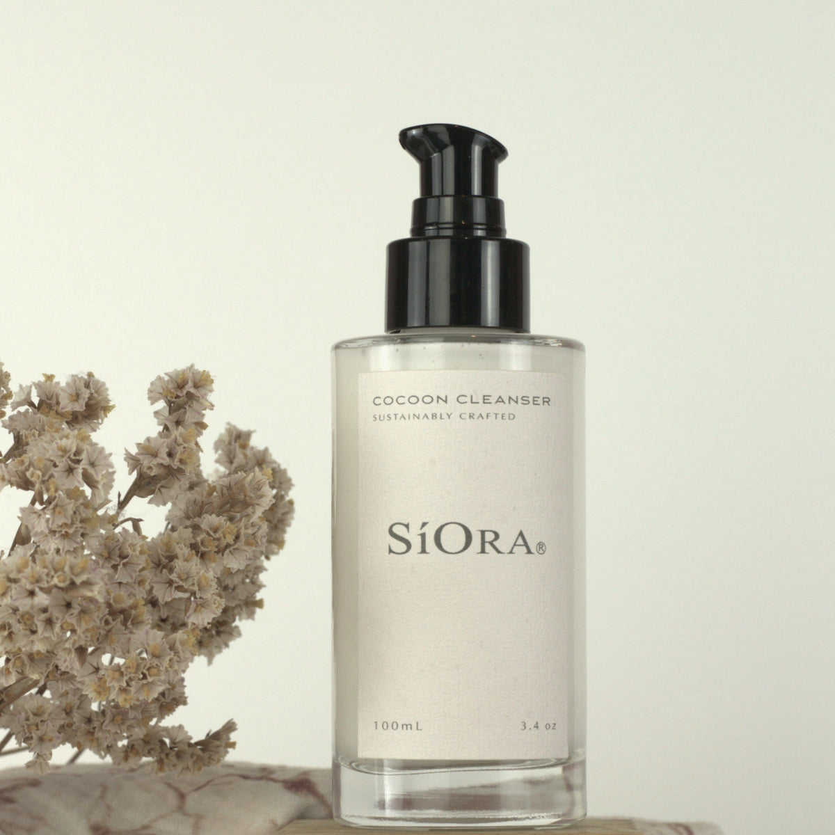 SiORA Cocoon Cleanser bottle with a black pump and clear glass body, displayed on a wooden block next to dried flowers and a patterned cloth against a warm cream background.