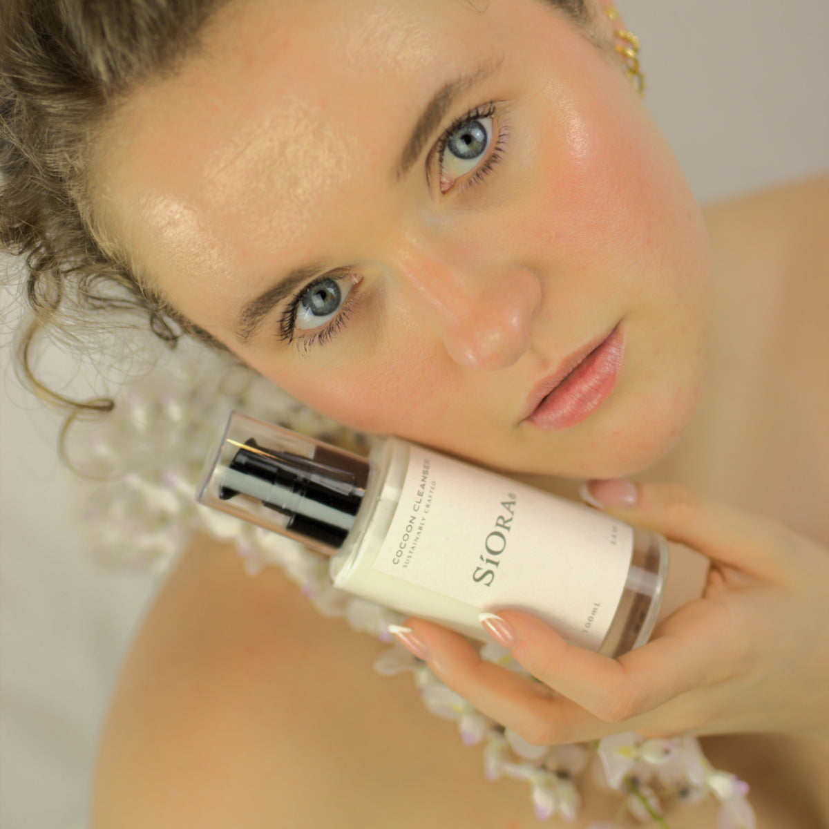 A woman with wet, curly brown hair holds a white bottle of Siora hair product near her face against a soft, light background.