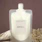 A stand-up pouch of SiORA Cocoon Cleaner natural coconut cleanser, filled with a milky white liquid, styled with a beige patterned cloth and dried flowers against a solid brown background.