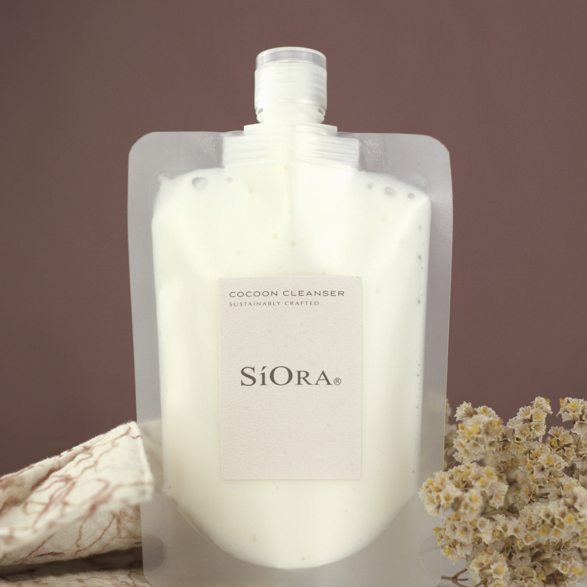 A stand-up pouch of SiORA Cocoon Cleaner natural coconut cleanser, filled with a milky white liquid, styled with a beige patterned cloth and dried flowers against a solid brown background.
