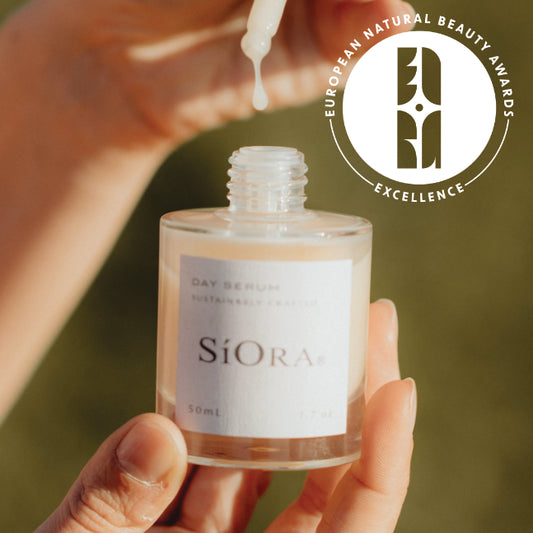 A hand holds a clear glass bottle of SiOra Day Serum, with another hand dripping the serum above it. The bottle has a white label with the brand name 'SiOra' and a 50ml size marking.