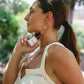 A woman in a cream-colored sleeveless top holds a small glass bottle with a black cap near her neck, set against a blurred outdoor background of green foliage and sunlight.