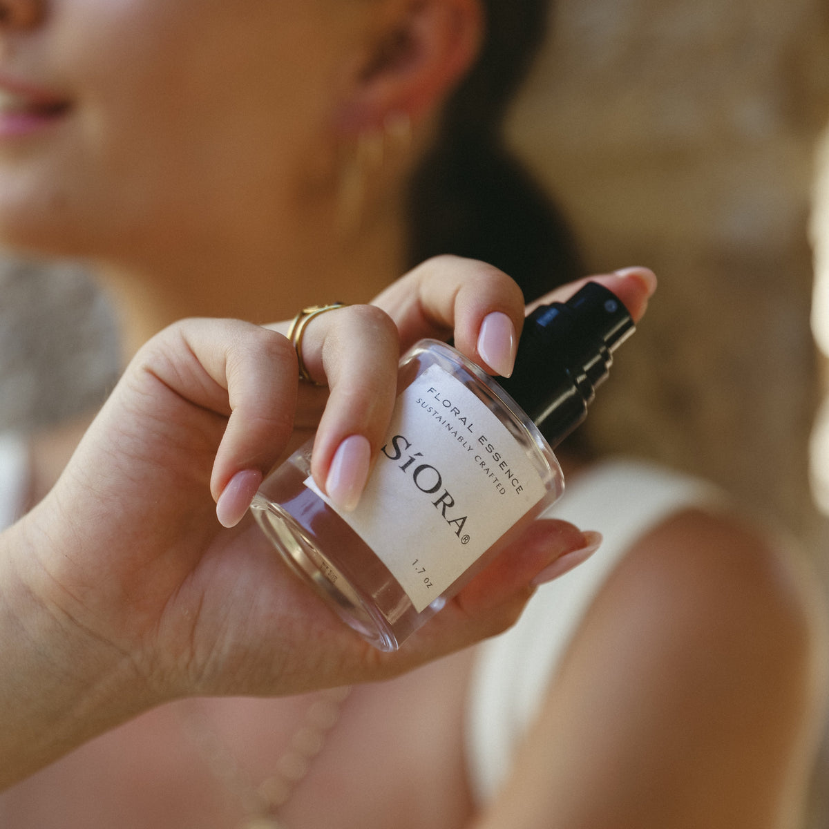 A woman holds a small clear glass perfume bottle labeled SIORA with a black spray top. The background is a warm, softly blurred stone wall and her face is out of focus, emphasizing the product.