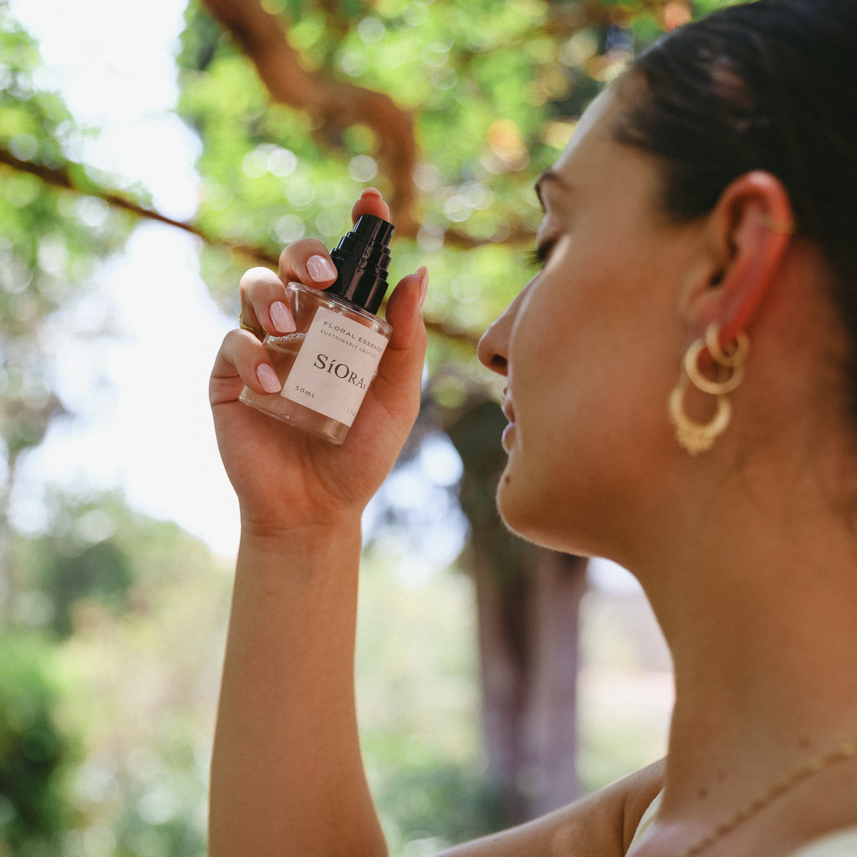 A woman in profile outdoors holds up a small SIORA cosmetic bottle to smell or inspect it. The bottle has a black dropper cap and a white label with the Dior name visible.