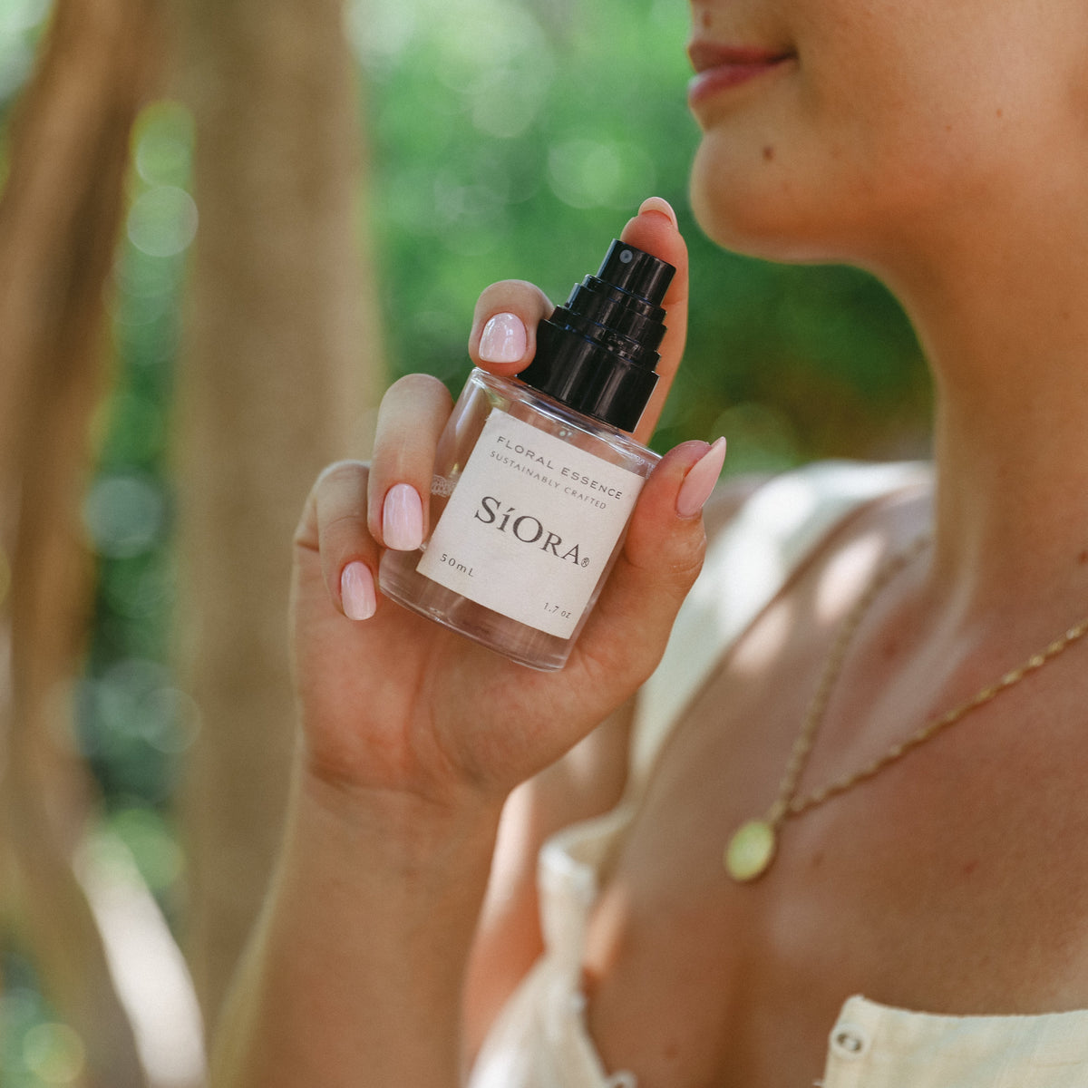 A woman outdoors holds a small clear bottle of Siora Floral Scents mist. The bottle has a black spray top and a white label with the brand name 'SIORA' printed in black.