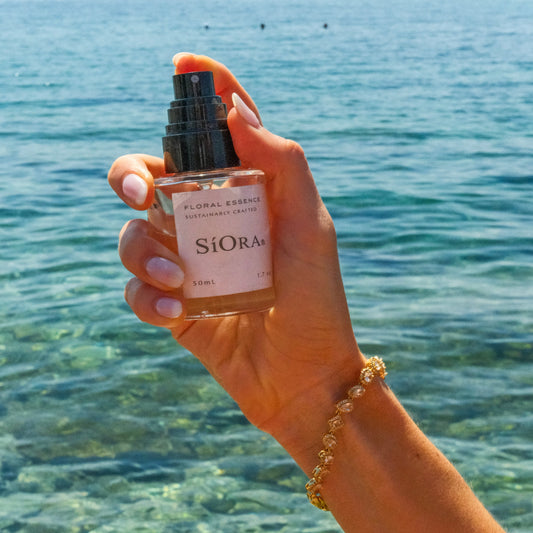 A hand holding a bottle of SiOra Floral Essence body mist against a scenic background of a clear blue sea and distant mountains.