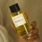 A hand holding a clear bottle of Siora luxury body oil with golden-yellow liquid and a black cap, set against a soft beige background.