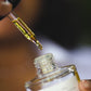 A close-up of a SIORA Luxury Oil dropper bottle with a black dropper cap, showing golden-yellow oil in the pipette above an open bottle. A hand is holding the dropper, and the background is softly blurred.
