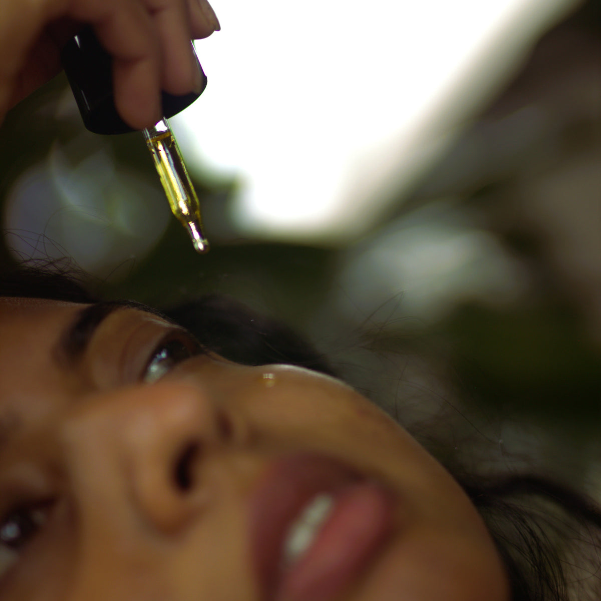 A close-up lifestyle shot of a person holding a dropper of SIORA luxury Oil bottle above their face, with a gold dropper containing golden oil hovering near their eye.